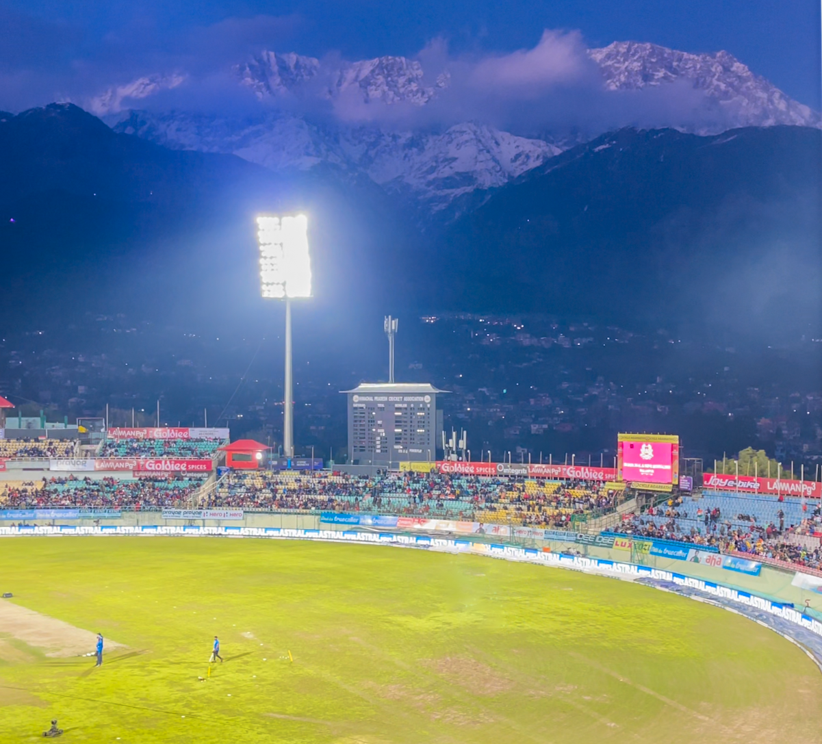 Evening match at the Dharamshala cricket stadium with floodlights and snow-capped Dhauladhar mountains