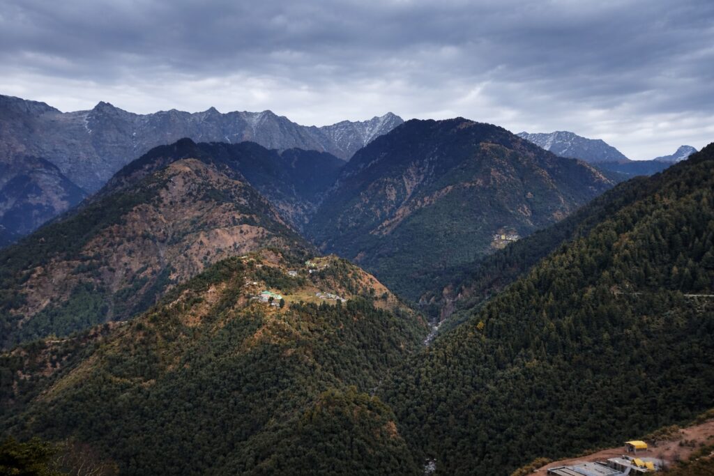 Dhauladhar mountain range in Dharamshala with forested hills, deep valleys, and snow-dusted Himalayan peaks under cloudy skies.