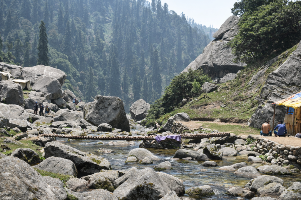 Mountain stream flowing through rocky valley with forested hills and hikers resting along a trekking trail near Dharamshala