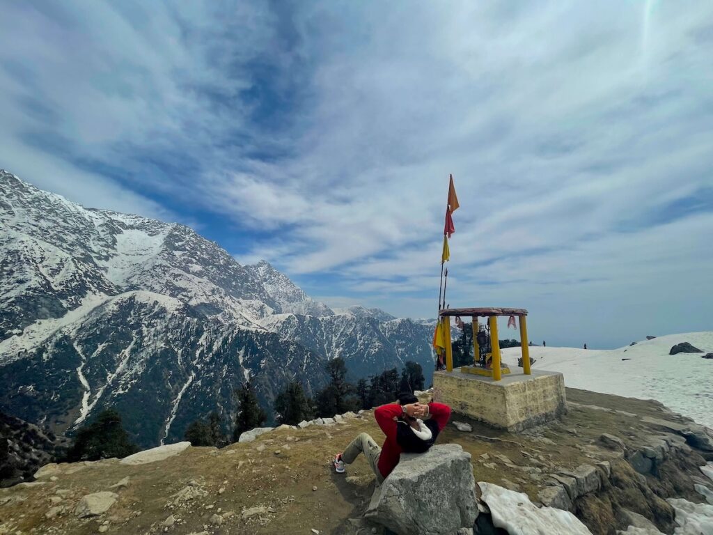 Hiker resting near a small shrine with snow-covered Dhauladhar mountains during trekking in Dharamshala