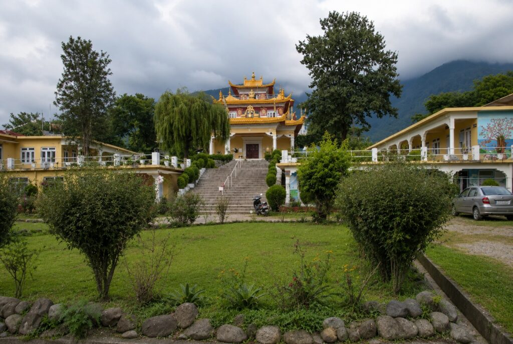 Bir Tibetan Colony monastery surrounded by lush gardens, trees, and mountain scenery under dramatic cloudy skies in Himachal Pradesh.