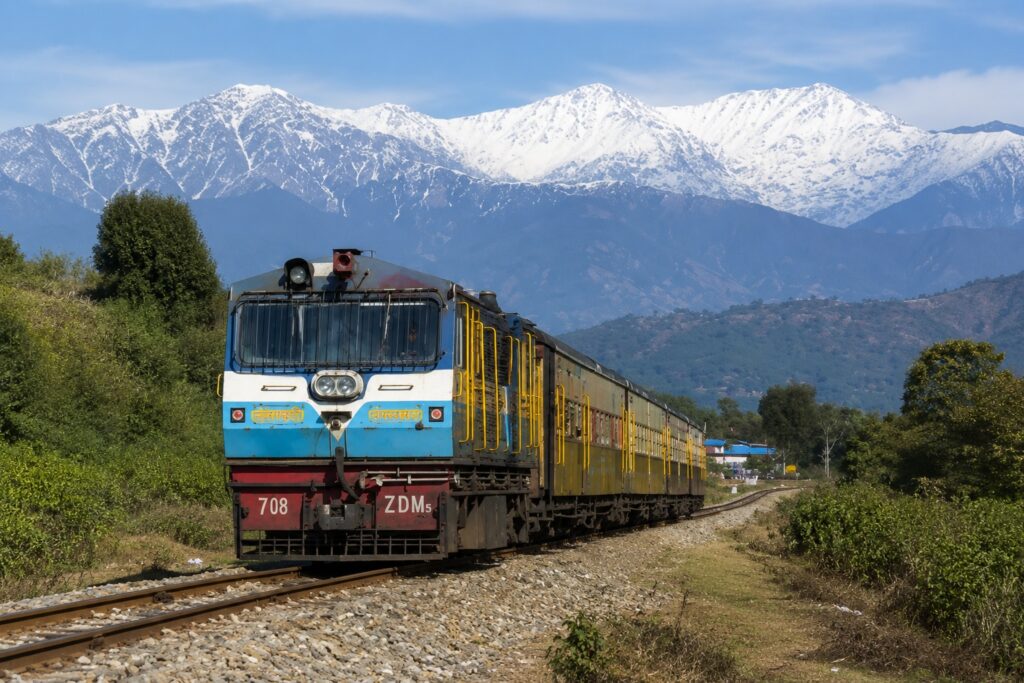 Kangra Valley Toy Train traveling through the foothills near Bir with snow-capped Dhauladhar mountains in the background, one of the most scenic things to do in Bir.