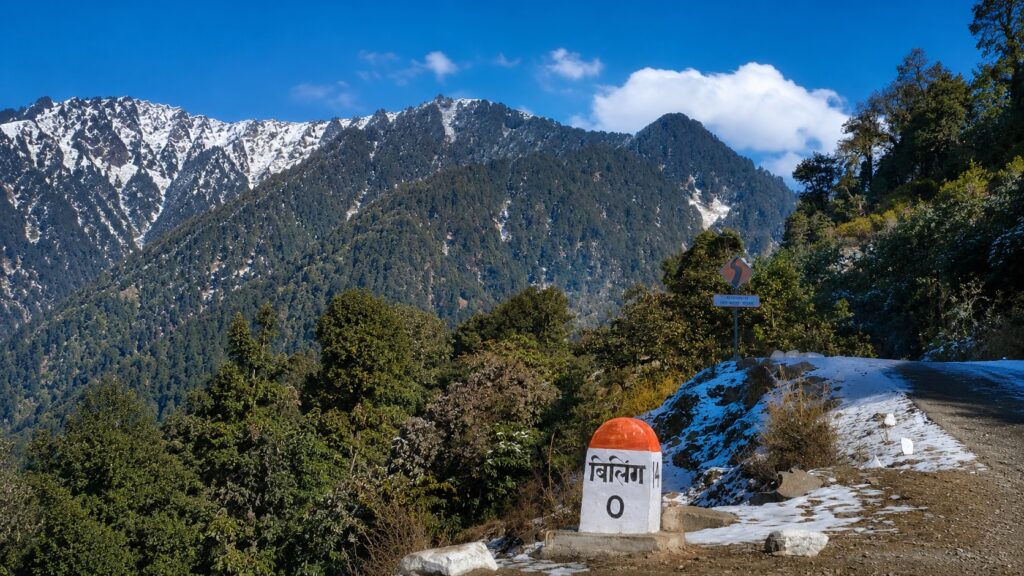Scenic mountain road leading to the Bir Billing paragliding takeoff point with snow-covered Himalayan peaks and the Billing milestone.