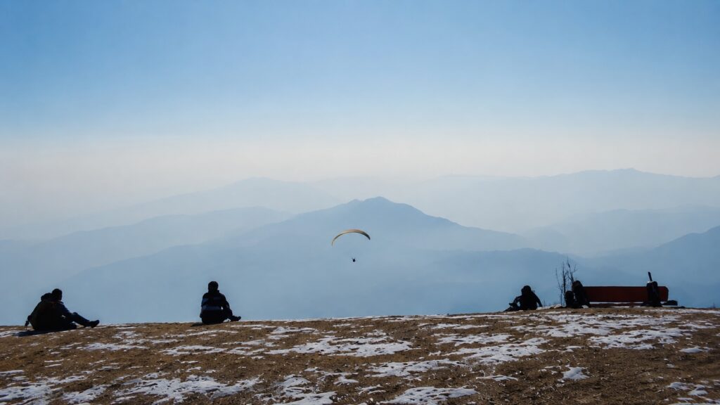 Bir Billing paragliding takeoff point with a paraglider soaring over misty Himalayan mountains and a snow-covered hillside in winter.